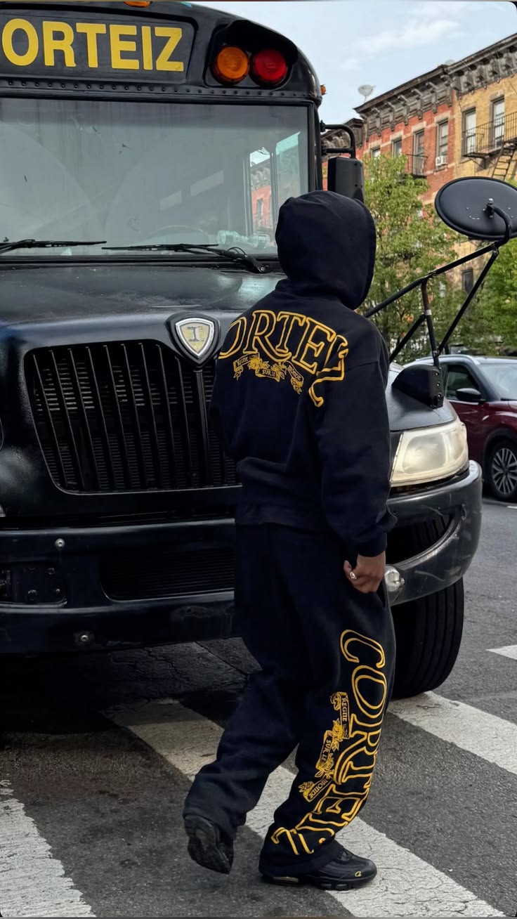 Person wearing a black hoodie and pants with 'DORTEIZ' branding standing next to a black bus.