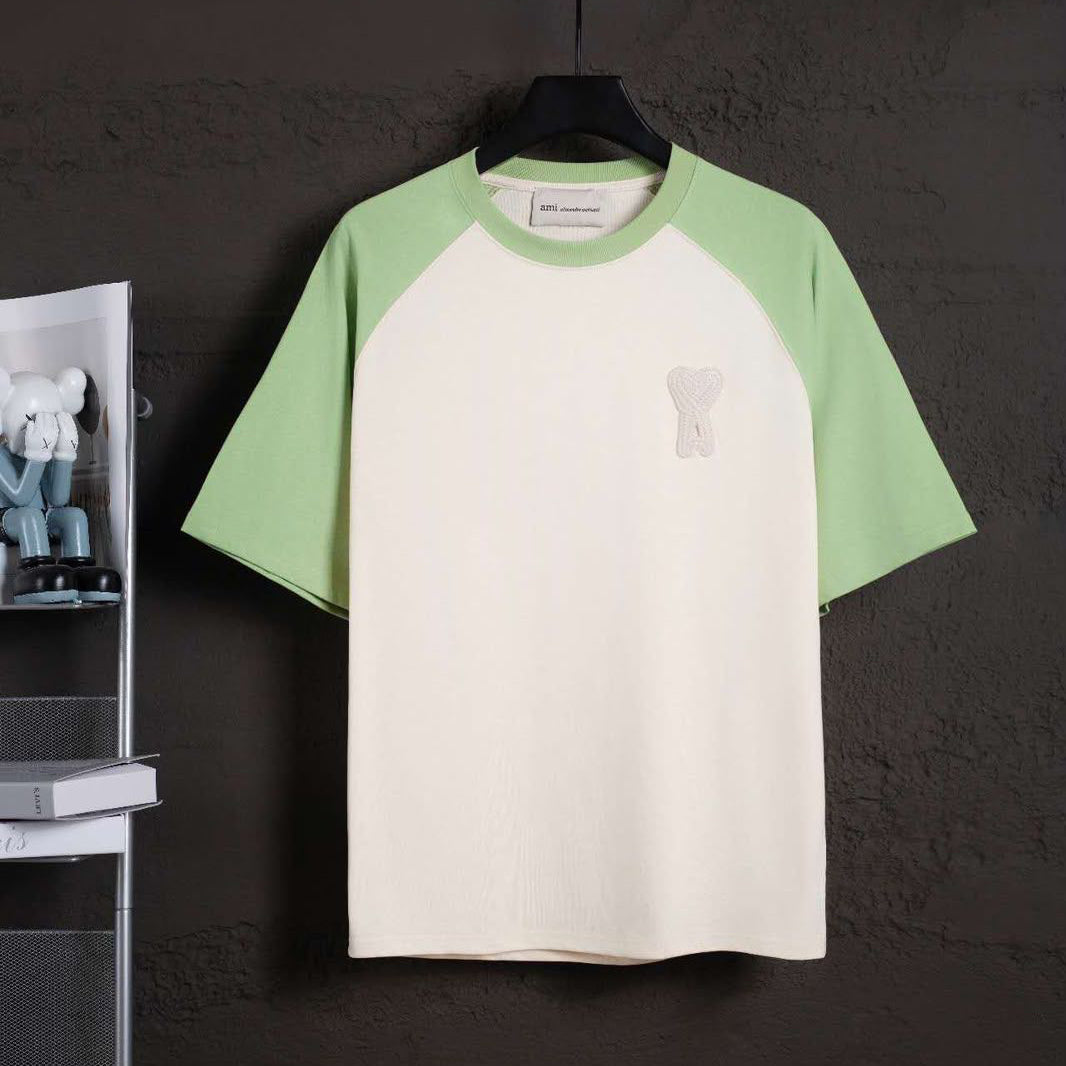 A short-sleeve raglan t-shirt hanging on a black hanger against a dark textured wall. The shirt features an off-white body and light green sleeves, with a small white embroidered graphic on the left chest.