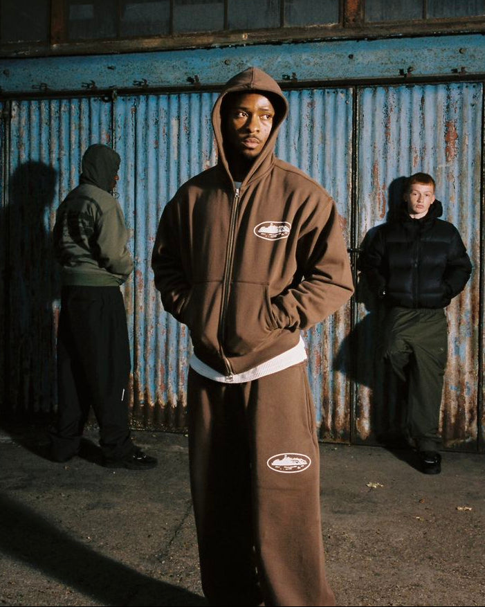 Person wearing a brown hoodie with a logo in front of a rusty metal wall.