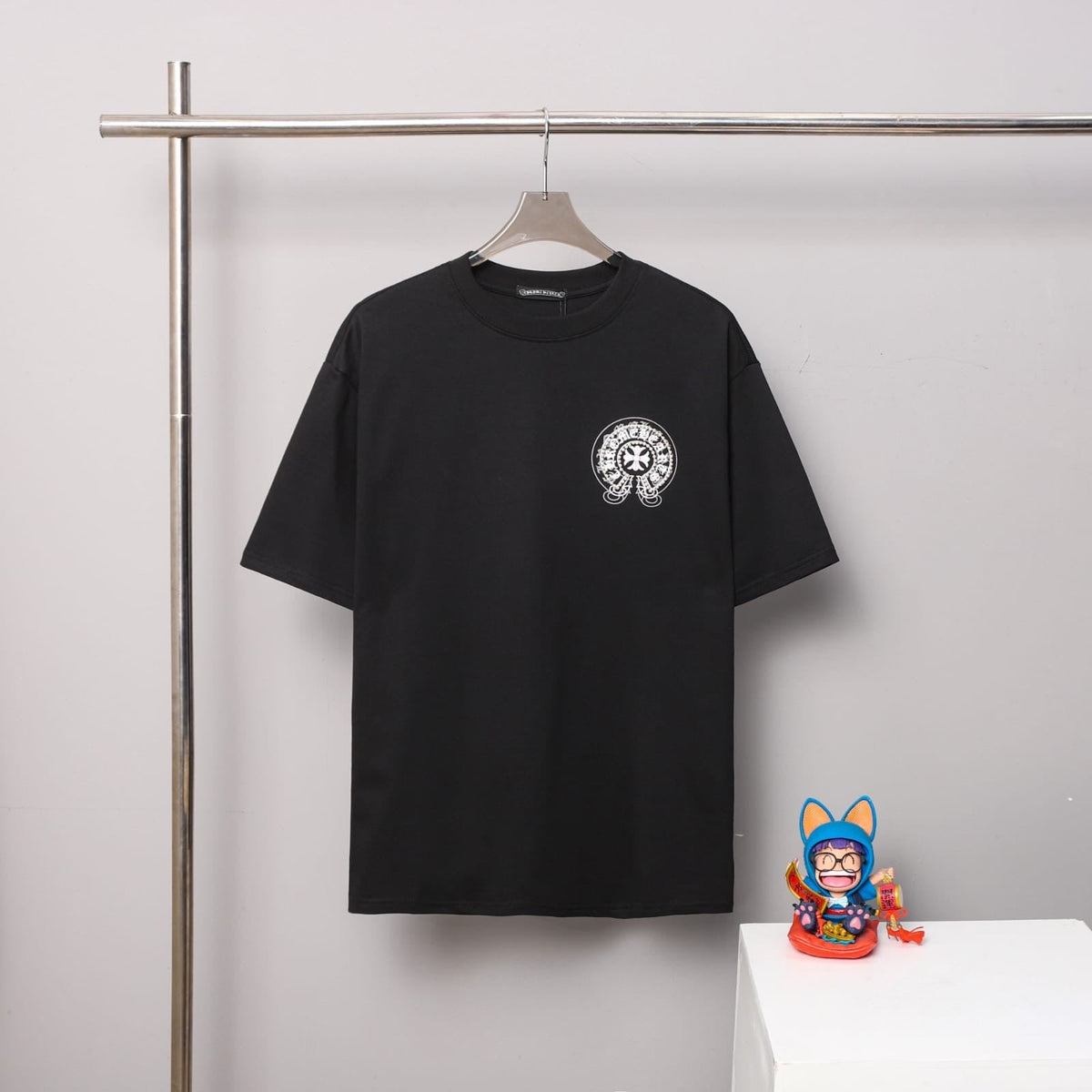 Black t-shirt with a logo on a hanger against a gray wall with a small figurine on a white surface.