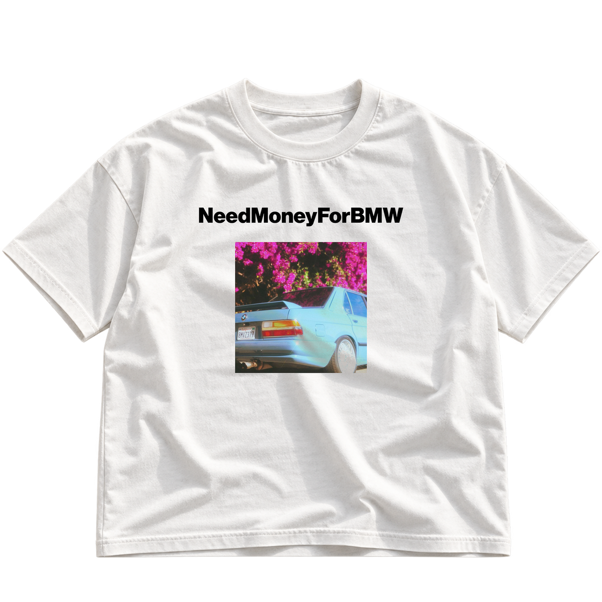 A white short-sleeve crewneck t-shirt featuring a graphic print on the front. The print displays the black text 'NeedMoneyForBMW' above a square image of a light blue BMW car parked under bright pink flowers.